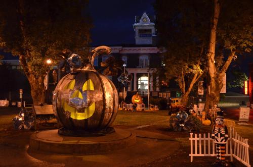 A large illuminated pumpkin sculpture surrounded by Halloween decorations at night, with a historic building in the background.