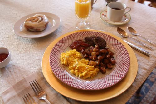 A breakfast plate with scrambled eggs, sausage, bacon, potatoes, a cinnamon roll, orange juice, and coffee.