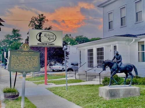 A statue of a horse rider in front of a building with a sign for Rebecca Ruth Bourbon Candy, under a colorful sunset.