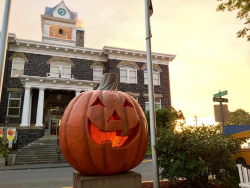 A large carved pumpkin with a smiling face stands in front of a historic city hall building at sunset.