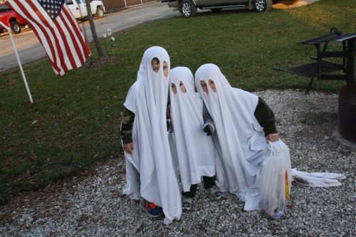 Three children dressed as ghosts in white sheets, standing on gravel with a flag in the background.
