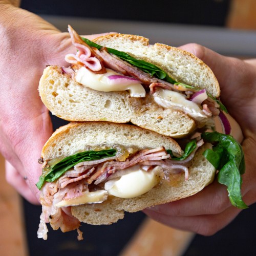 A close-up of a hand holding a sandwich filled with ham, cheese, spinach, and onions, showcasing fresh ingredients.