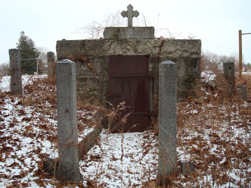 A weathered stone mausoleum with a cross on top, surrounded by snow and overgrown vegetation.