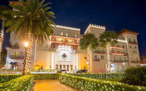 A beautifully lit hotel at night, surrounded by palm trees and festive lights, creating a warm, inviting atmosphere.