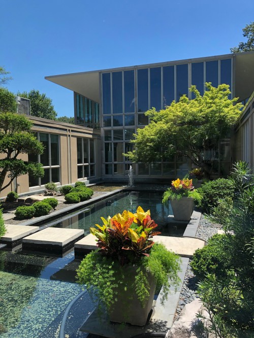 A modern garden with a reflecting pool, vibrant plants, and a contemporary building under a clear blue sky.