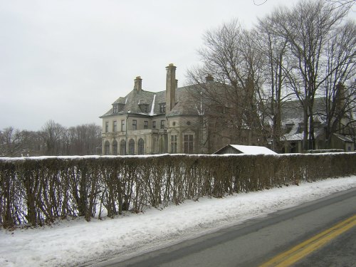 A large, historic mansion surrounded by snow and bare trees, viewed from a road on a cloudy winter day.