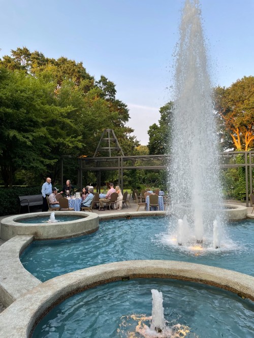 A serene garden scene featuring a fountain, surrounded by greenery and people enjoying a gathering at tables.