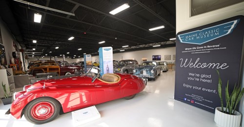 A red classic car in a museum with various vintage cars in the background and a welcome sign nearby.