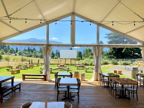 A spacious tent interior with wooden tables, overlooking a scenic outdoor area with mountains and picnic tables.