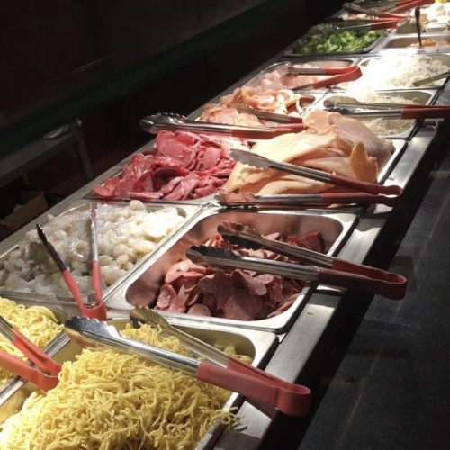 A variety of fresh ingredients displayed in trays, including meats, noodles, and vegetables, with tongs for serving.