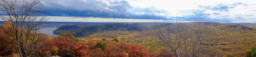 Panoramic view of colorful autumn foliage covering rolling hills and a river under a cloudy sky.