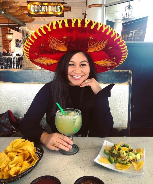 A woman wearing a large sombrero smiles while holding a cocktail, with nachos and guacamole on the table in front of her.
