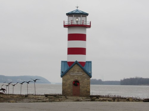 A red and white striped lighthouse stands by a river, with a stone base and a cloudy sky in the background.
