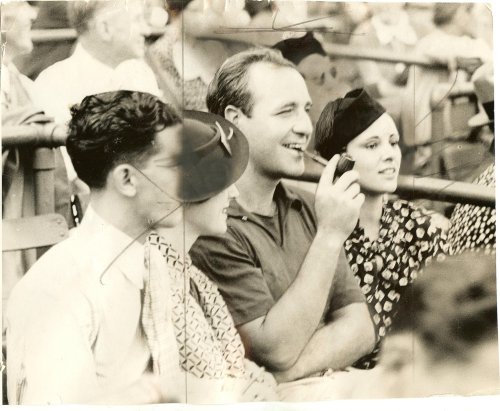 A vintage photo of three people sitting together, smiling and enjoying an event, with a crowd in the background.