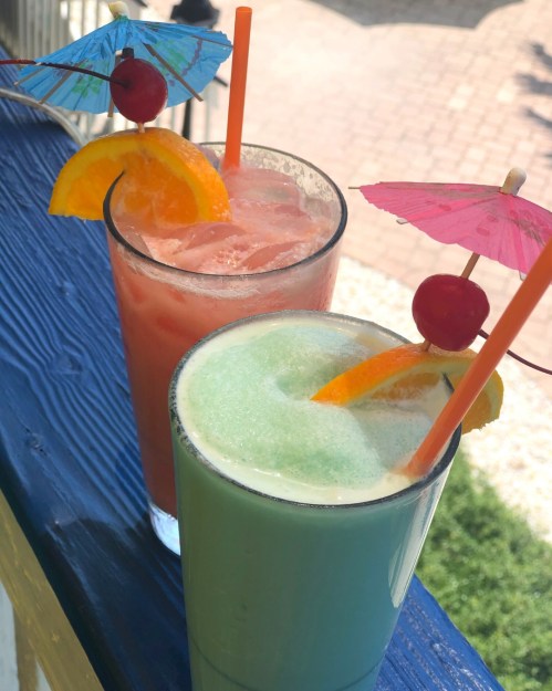Two colorful cocktails with umbrellas, garnished with orange slices and cherries, on a blue railing.