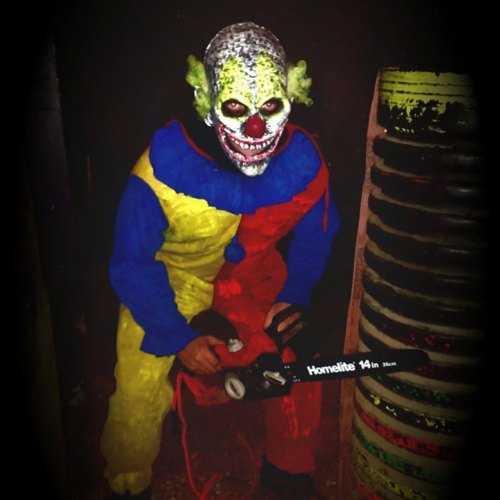 A creepy clown in a colorful costume holds a chainsaw, standing in a dimly lit environment.