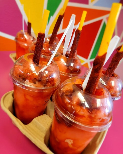 Colorful drinks in clear cups with tamarind sticks, served in a cardboard tray against a vibrant background.