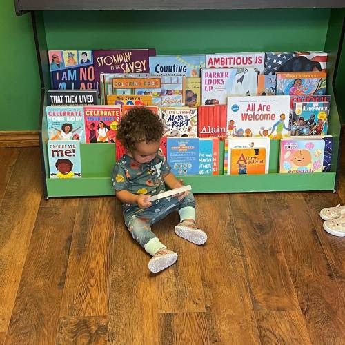 A young child sits on the floor, reading a book in front of a colorful bookshelf filled with children's books.