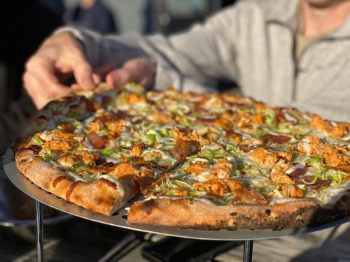 A person holds a slice of pizza topped with chicken, green peppers, and cheese, on a metal tray outdoors.
