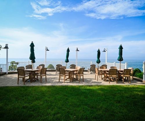 Outdoor patio with wooden tables and chairs, green umbrellas, overlooking a calm sea under a blue sky.