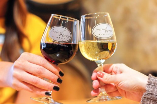 Two hands clink wine glasses, one filled with red wine and the other with white, both labeled Prairie Berry Winery.