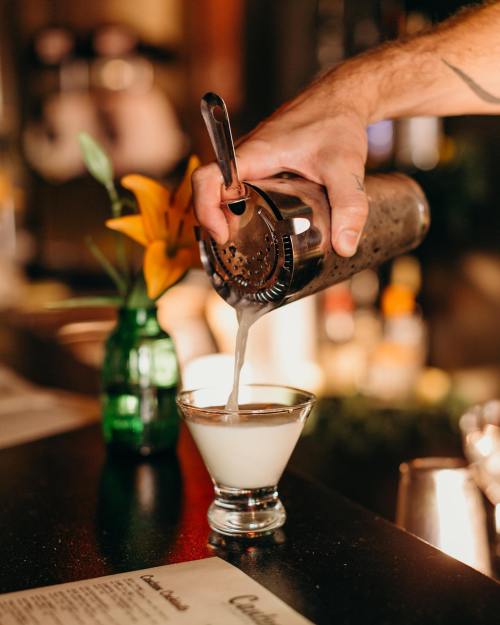 A hand pours a cocktail from a shaker into a glass, with a flower in a vase and a menu in the background.