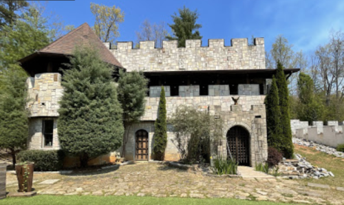 A stone castle-like building surrounded by trees and a landscaped garden under a clear blue sky.