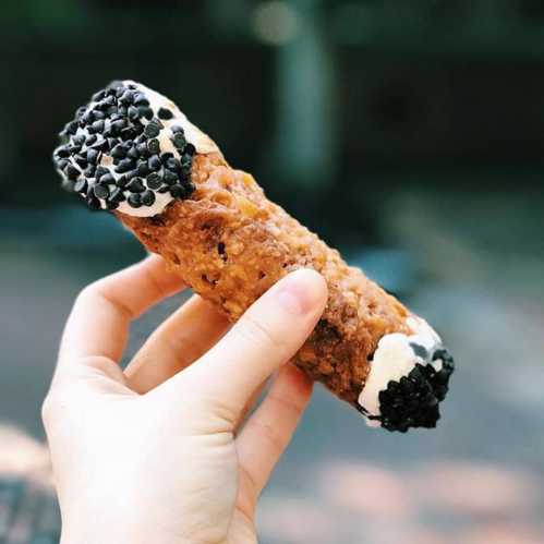 A hand holding a cannoli topped with chocolate sprinkles and cream, set against a blurred outdoor background.