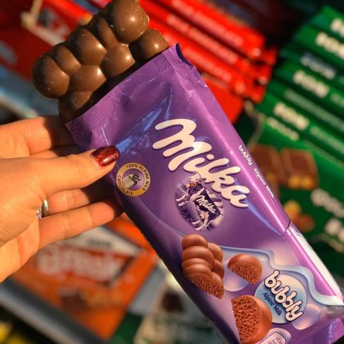 A hand holding a bar of Milka Bubbly chocolate, featuring a purple wrapper with chocolate pieces visible.