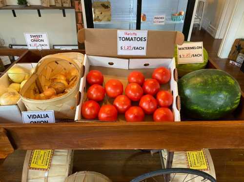 A wooden cart displays fresh produce: tomatoes, sweet onions, Vidalia onions, and a watermelon, with price tags.