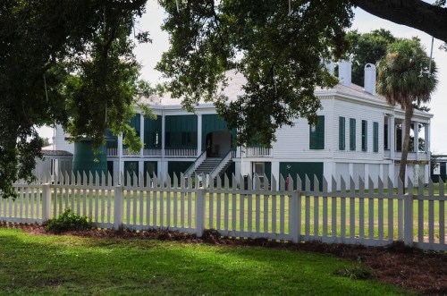 A historic white house with green shutters, surrounded by a white picket fence and lush greenery.