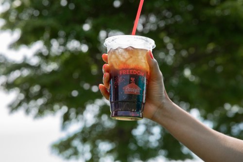A hand holding a clear cup of iced coffee with a red straw, set against a blurred green background.