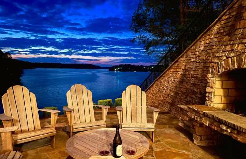 A serene lakeside view at dusk, featuring wooden chairs, a table with wine, and a stone wall.