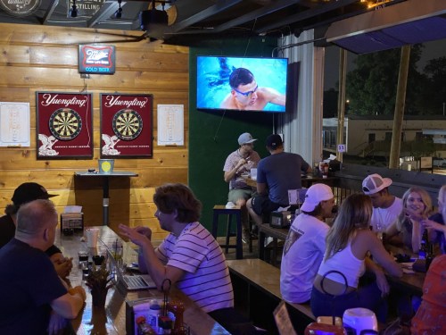 A lively bar scene with patrons at tables, dartboards on the wall, and a TV showing a swimming event.