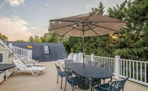 A spacious outdoor patio with a table, chairs, and an umbrella, surrounded by greenery and a clear sky.