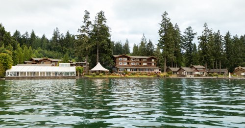 A serene lakeside view featuring a lodge and several cabins surrounded by tall trees under a cloudy sky.