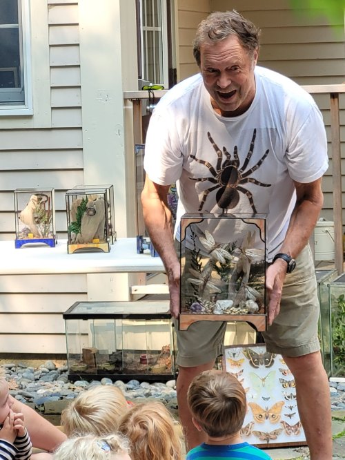 A man in a spider-themed shirt holds a display case while children watch attentively outdoors.