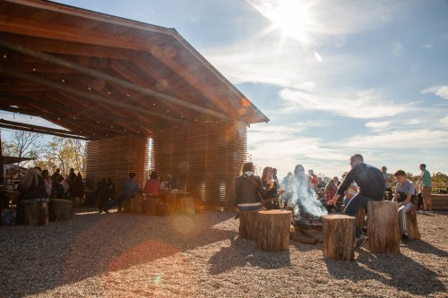 A gathering of people around a fire pit outside a wooden structure, with sunlight shining and a clear blue sky.