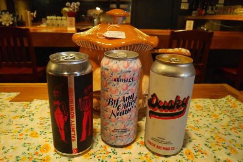 Three beer cans on a table, featuring colorful labels, with a decorative jar in the background.