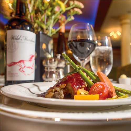 A gourmet dish with meat, garnished with asparagus and cherry tomatoes, alongside a glass of red wine and floral decor.