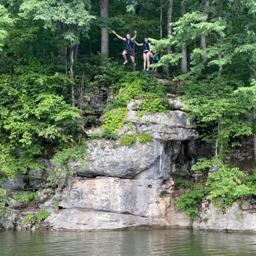 Two people stand on a rocky ledge by a lake, surrounded by lush green trees, ready to jump into the water.