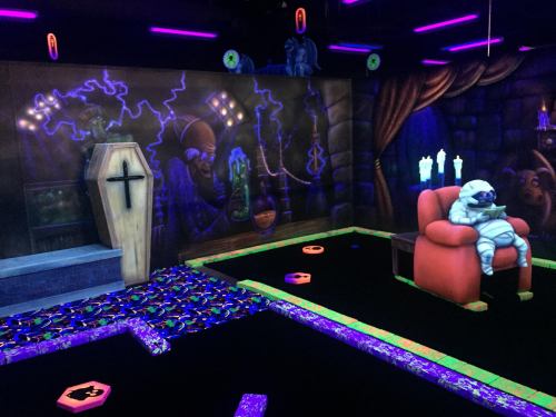 A spooky mini-golf scene featuring a coffin, eerie decorations, and a ghostly figure sitting in a red chair.