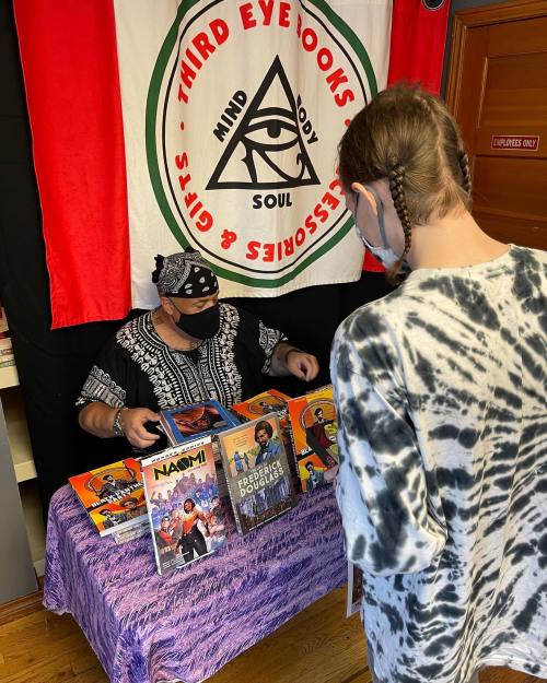 A person browses comic books at a table, while another person, wearing a mask, assists them. A colorful banner is in the background.