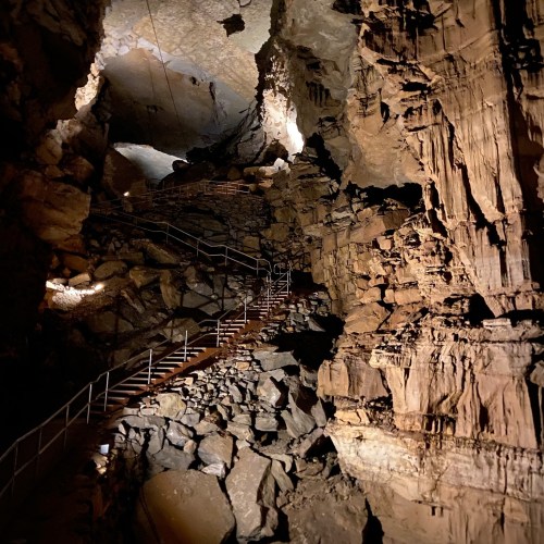 A dimly lit cave interior with rocky walls and a winding metal staircase leading deeper into the cavern.