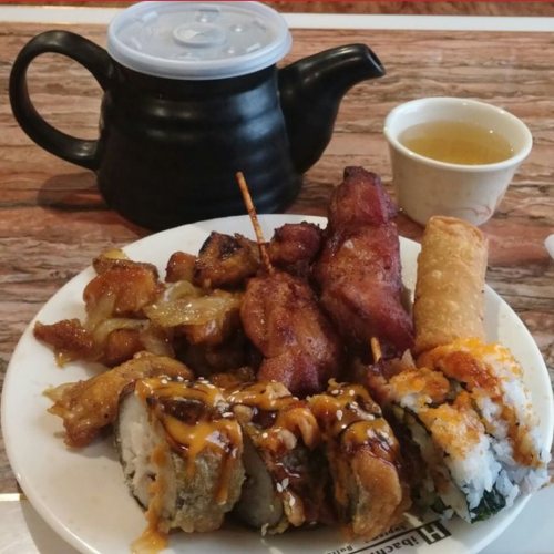 A plate of sushi rolls, fried chicken, and a spring roll, with a small teapot and cup in the background.