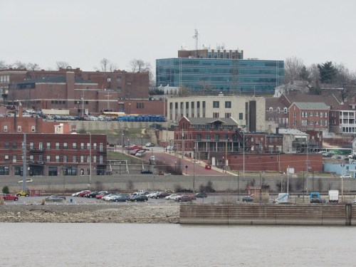 A view of a riverside cityscape featuring buildings, parking lots, and a cloudy sky.
