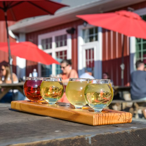 A wooden flight paddle with four colorful drinks sits on a table, with red umbrellas and people in the background.
