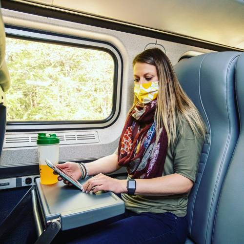 A woman in a mask sits on a train, using her phone and sipping a drink, with trees visible through the window.