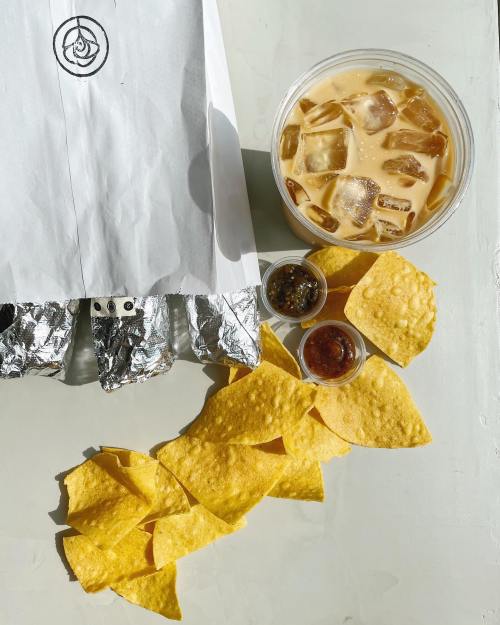 A takeout bag next to a cup of iced drink, with tortilla chips and two small containers of salsa on a light surface.