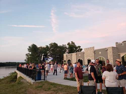 A lively outdoor event by a lake, with a crowd gathered near a stage and a building, under a colorful sunset sky.